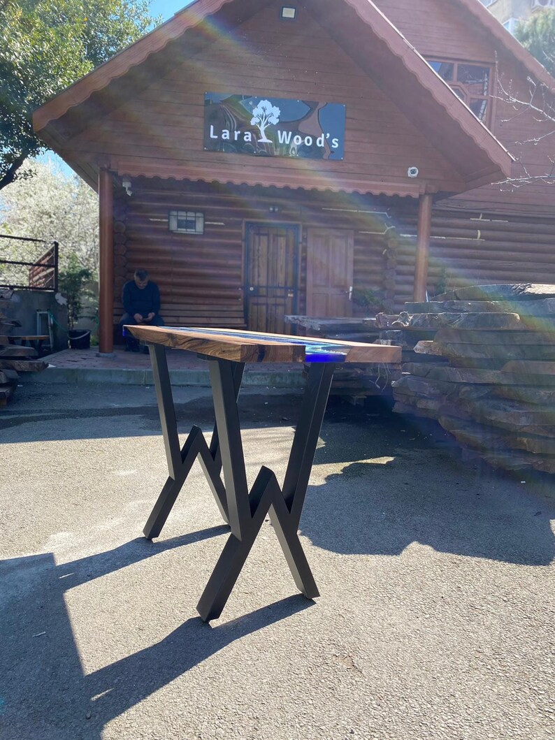 May include: A handcrafted wooden table with a dark wood top and blue resin accents. The table has black metal legs with a unique zigzag design. The business name "Lara Wood's" is visible in the background.