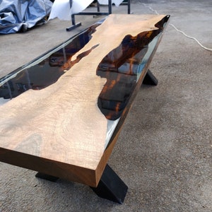 May include: A wooden bench with a live edge design. The tabletop is light-coloured wood with dark, glossy epoxy resin filling the natural voids. The bench has black metal legs, creating a modern, industrial aesthetic. The bench is approximately 150 cm long.