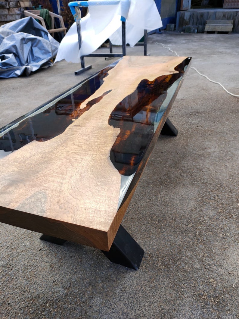 May include: A wooden bench with a live edge design. The tabletop is light-coloured wood with dark, glossy epoxy resin filling the natural voids. The bench has black metal legs, creating a modern, industrial aesthetic. The bench is approximately 150 cm long.