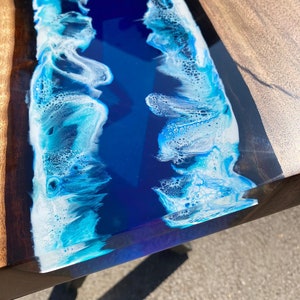 May include: A close-up of a wooden table with a river of blue resin. The resin is a deep blue color with white foamy edges, resembling ocean waves. The wooden sides are a light brown color.