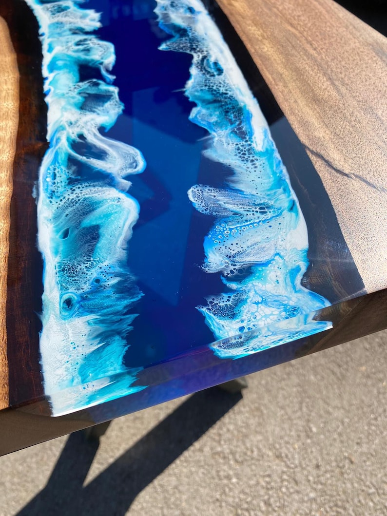 May include: A close-up of a wooden table with a river of blue resin. The resin is a deep blue color with white foamy edges, resembling ocean waves. The wooden sides are a light brown color.