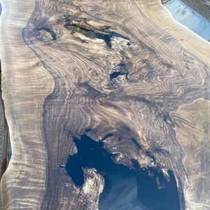 May include: A large, live edge wooden table with a dark, glossy finish. The wood grain is prominent, with areas of dark brown and lighter tan. The table has several dark, resin-filled voids, adding a unique design element. The table's dimensions are not visible.