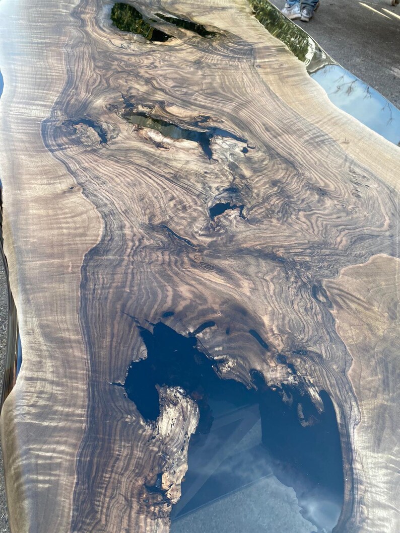 May include: A large, live edge wooden table with a dark, glossy finish. The wood grain is prominent, with areas of dark brown and lighter tan. The table has several dark, resin-filled voids, adding a unique design element. The table's dimensions are not visible.