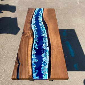 May include: A rectangular wooden coffee table with a blue epoxy resin river design. The table has a dark brown wood grain finish on the sides and a vibrant blue and white river design in the center.