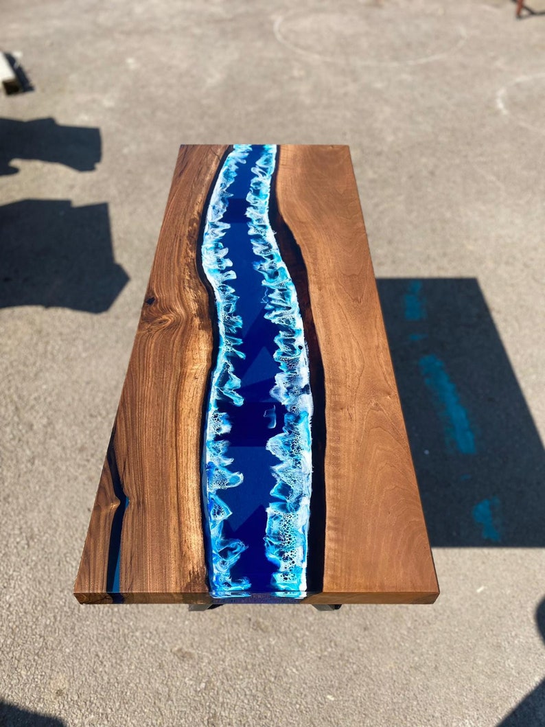May include: A rectangular wooden coffee table with a blue epoxy resin river design. The table has a dark brown wood grain finish on the sides and a vibrant blue and white river design in the center.