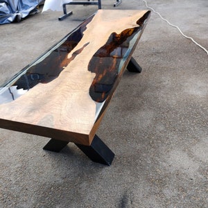 May include: A handcrafted wooden table with a live edge and a dark epoxy resin river. The table has a light brown wood surface with dark brown and black accents. It is supported by black metal legs. The table measures approximately 183 cm long.