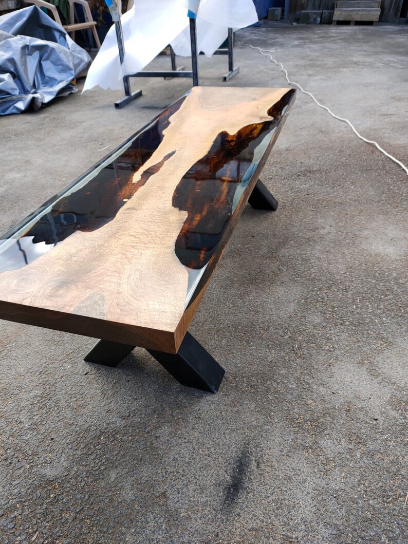 May include: A handcrafted wooden table with a live edge and a dark epoxy resin river. The table has a light brown wood surface with dark brown and black accents. It is supported by black metal legs. The table measures approximately 183 cm long.