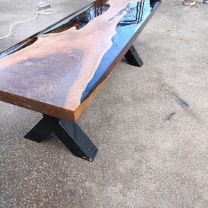 May include: A handcrafted wooden table with a live edge design. The tabletop features a dark wood grain with a clear epoxy resin river. The table has black metal legs in an X-shape. The table is approximately 183 cm long.