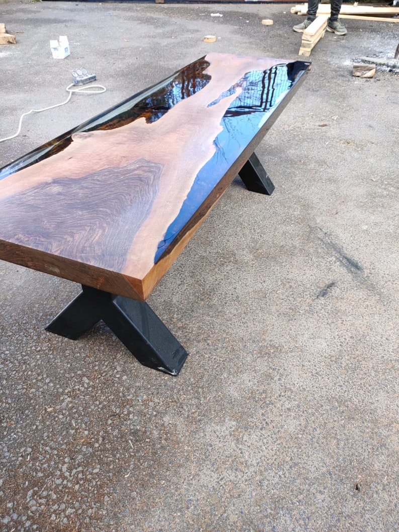 May include: A handcrafted wooden table with a live edge design. The tabletop features a dark wood grain with a clear epoxy resin river. The table has black metal legs in an X-shape. The table is approximately 183 cm long.
