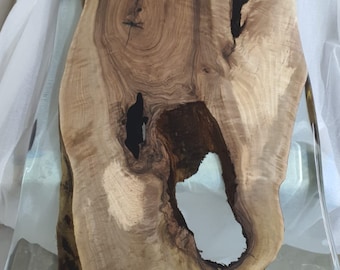 Puede incluir: Una mesa de madera con acabado de resina transparente. La mesa presenta un patrón de vetas de madera natural con nudos oscuros y una gran abertura irregular. Los bordes son lisos y la resina proporciona una superficie brillante.