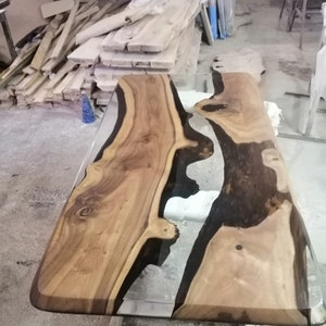 Puede incluir: Mesa de madera artesanal con un centro de resina epoxi transparente. La mesa presenta un patrón de vetas de madera natural con tonos marrones claros y oscuros. Los bordes son redondeados y la mesa está soportada por un marco metálico.