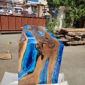 May include: A handcrafted wooden table with a unique design. The table features a rich, brown wood grain interspersed with vibrant blue resin, creating a striking contrast. The table's dimensions are approximately 122 cm long, 61 cm wide, and 76 cm high.