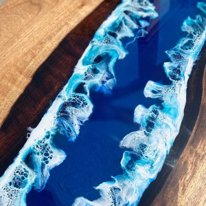 May include: Close-up of a wooden table with a river of blue resin. The resin has white and light blue wave-like patterns. The wood has a natural grain and a warm, brown color.