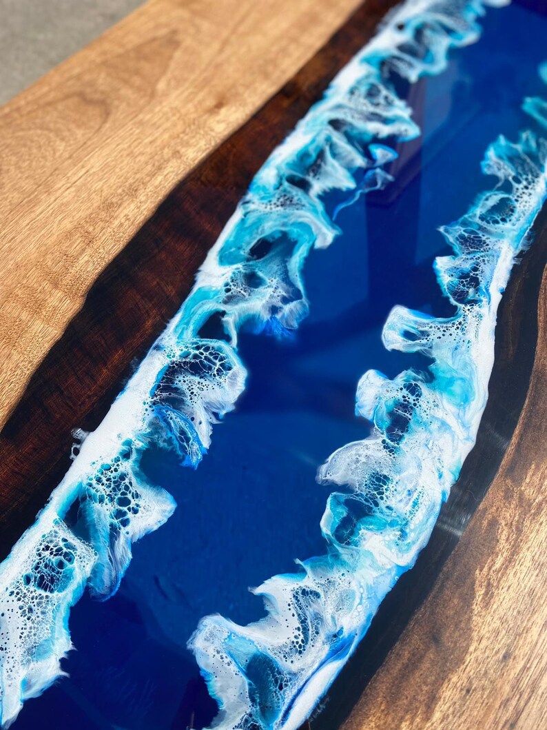 May include: Close-up of a wooden table with a river of blue resin. The resin has white and light blue wave-like patterns. The wood has a natural grain and a warm, brown color.