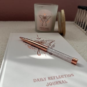 May include: A rose gold pen with a crystal barrel lies on a white journal with the words "DAILY REFLECTION JOURNAL" printed in pink. A candle with a butterfly design and the words "WHITE SAGE" is in the background.