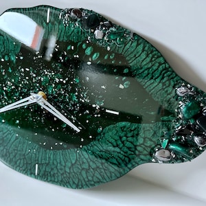 Peut inclure: Une horloge en résine verte avec des accents argentés et verts. Le cadran de l'horloge est d'un vert foncé avec des aiguilles blanches et un centre doré. L'horloge a la forme d'un ovale libre.