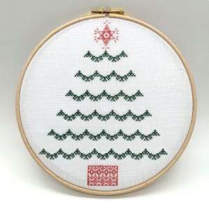 Puede incluir: Un bastidor de bordado de punto de cruz con un diseño de árbol de Navidad. El árbol está compuesto por puntadas verdes, con una estrella roja en la parte superior y un regalo rojo en la base. El bordado está en un bastidor de madera.