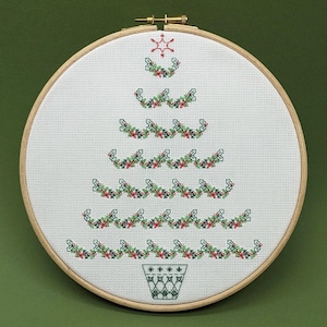 Puede incluir: Un aro de bordado blanco con un diseño de punto de cruz de un árbol de Navidad. El árbol está hecho de guirnaldas verdes con bayas rojas y una estrella en la parte superior. El árbol está en una maceta verde con un diseño de copo de nieve blanco.