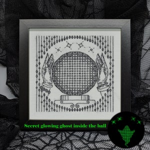 May include: A black and white cross-stitch design of a crystal ball held by two hands. The design is framed in a black frame. The text "Secret glowing ghost inside the ball" is below the frame. A green glowing ghost is in the lower right corner of the image.