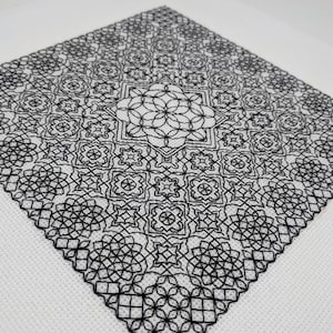 Puede incluir: Bordado de punto de cruz en blanco y negro con un patrón geométrico. El diseño presenta un motivo floral central rodeado de formas repetidas y detalles intrincados. La pieza está sobre un fondo de tela blanca.