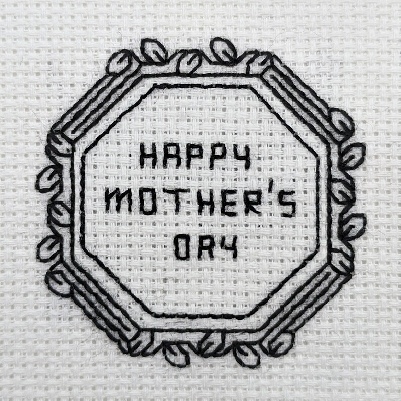 Pode incluir: Tecido branco com bordado preto de um oct&oacute;gono com o texto "Happy Mother's Day" dentro.