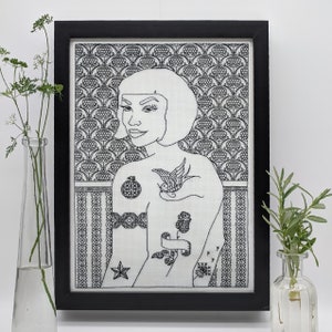 May include: Black and white embroidery of a woman with a short haircut and tattoos on her arms and chest. The tattoos include a bird, a flower, and a heart. The woman is framed by a black frame and a patterned background.