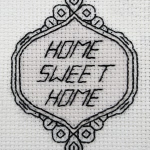 Pode incluir: Desenho bordado em preto e branco sobre um fundo de tecido branco. O desenho apresenta uma moldura decorativa com o texto "HOME SWEET HOME" no seu interior.