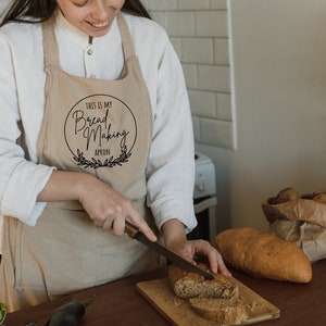 K&ouml;nnte beinhalten: Eine Person tr&auml;gt eine beige Sch&uuml;rze mit dem Text "This is my Bread Making Apron" in einem Kreis mit einem Kranz drumherum. Die Person schneidet ein Brot auf einem Holzbrett.