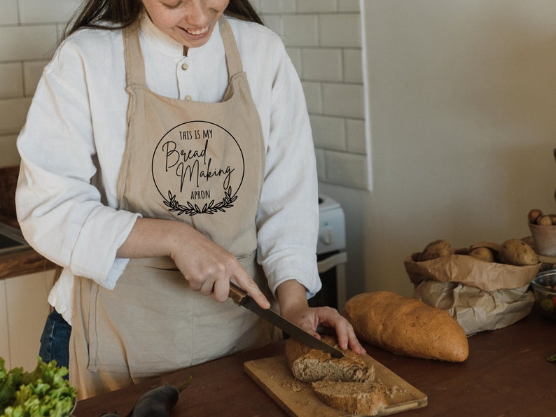 K&ouml;nnte beinhalten: Eine Person tr&auml;gt eine beige Sch&uuml;rze mit dem Text "This is my Bread Making Apron" in einem Kreis mit einem Kranz drumherum. Die Person schneidet ein Brot auf einem Holzbrett.
