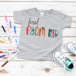May include: A gray t-shirt with the text "feral farm kid" in colorful letters with farm-related icons. The shirt is laid flat on a white wooden surface with markers, a potted plant, and a pair of light green sneakers.