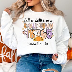 May include: White sweatshirt with the text "fall is better in a SMALL TOWN 98765 nashville, tn" in orange, purple, and yellow. The design includes a pumpkin and sunflowers. The sweatshirt is a fall-themed item.