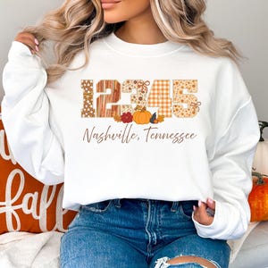 May include: White sweatshirt with the numbers "12345" in a patchwork design, with pumpkins and leaves. The text "Nashville, Tennessee" is below the numbers. The sweatshirt is worn by a person wearing blue jeans.