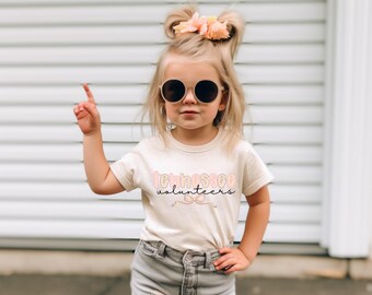 Op de afbeelding: Een jong kind draagt een crèmekleurig T-shirt met de woorden "Tennessee Volunteers" in roze, perzik en zwarte letters. Het kind draagt ook een zonnebril en een grijze spijkerbroek.