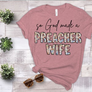 May include: A pink t-shirt with the text "So God made a Preacher Wife" in black lettering. The word "Preacher" is decorated with a floral pattern.