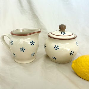 May include: Cream-colored ceramic creamer and sugar bowl set with a brown rim and handle. The set is decorated with blue floral patterns. The sugar bowl has a matching lid with a wooden knob. A yellow lemon is in the background.