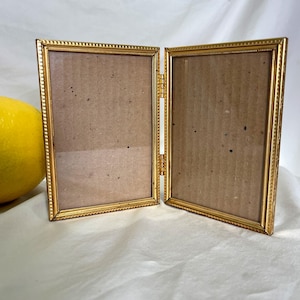 May include: A gold-colored, hinged double picture frame. The frame has a decorative beaded edge and contains two rectangular openings with brown backing. The frame is open, revealing the hinge and the backing material.