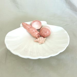 May include: A white ceramic seashell-shaped dish with a collection of pink ceramic seashells. The dish has a textured surface and contains four different seashell designs in varying shades of pink.
