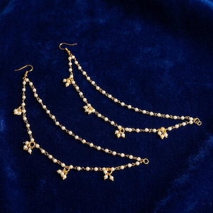 May include: A pair of gold earrings with white pearl detailing. The earrings have a chain design with multiple pearl strands and small pearl clusters. The background is a deep blue colour.