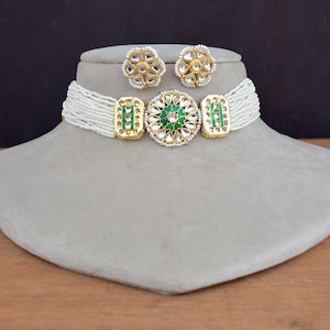 以下が含まれることがあります： 緑と金のペンダントとマッチするイヤリングが付いた白い真珠のチョーカーネックレス。ペンダントは、中央に小さな緑色の石がついた大きな丸いデザインです。