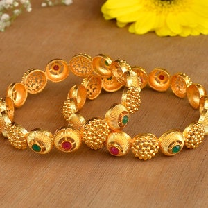 May include: Two gold bangles with intricate designs and green and red gemstones.