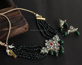 May include: A multi-strand necklace with green beads and a large, ornate pendant featuring a red stone surrounded by clear stones. The pendant is accented with green and white stones. Matching earrings with green and white stones are also included.