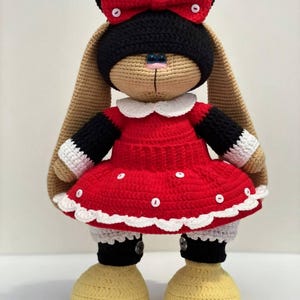 May include: A handmade crochet rabbit doll featuring long tan ears, a black head, and a red dress with white polka dots and a white collar. The doll wears a large red bow with white buttons, black sleeves with white cuffs, and large yellow shoes.