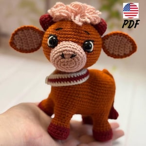 May include: A crocheted brown and pink cow plush toy with a red and white scarf. The toy has a pink nose and black eyes.