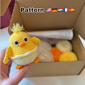 Puede incluir: Un pollito amarillo tejido a crochet con detalles blancos, sentado en una caja de cartón marrón con la palabra "Pattern" y las banderas de Estados Unidos, Alemania, Países Bajos, Francia y España.