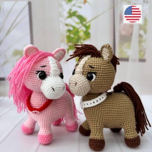 Może przedstawiać: Dwie ręcznie robione figurki koni na szydełku. Jeden jest różowy z białą twarzą i różową grzywą, drugi jest brązowy z białą twarzą i brązową grzywą. Oba mają czarne oczy i noszą czerwony lub biały kołnierz.