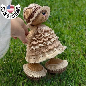 Pode incluir: Uma boneca de gato de crochê vestindo um vestido marrom com detalhes em renda e um chapéu combinando. A boneca está em pé em uma superfície de grama verde.
