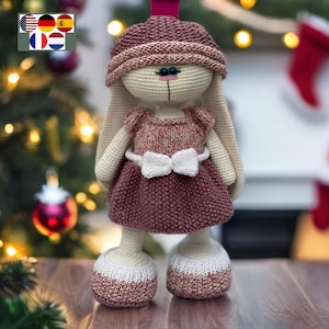 以下が含まれることがあります： ピンクと白のドレスを着た、白いリボンをつけた、かぎ針編みのウサギのぬいぐるみ。人形は青い目をしており、木製の表面に立っています。