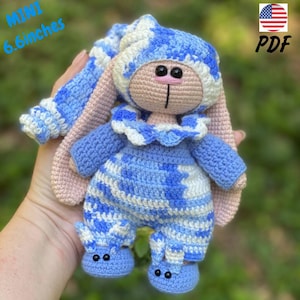 Op de afbeelding: Een gehaakte konijnenpop, ongeveer 17 cm hoog, gekleed in een blauw-wit patroon outfit met een bijpassende hoed en slofjes. Het konijn heeft roze oren en een bruin gezicht met zwarte ogen en een roze neus. De tekst "MINI 6.6 inches" en "PDF" is inbegrepen.