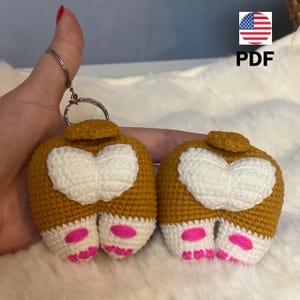 May include: Two crocheted dog bum keyrings. The keyrings are brown with white hearts and pink paw prints. The keyrings are attached to a silver keyring.