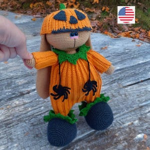 Pode incluir: Um coelho de pelúcia de crochê vestindo uma fantasia de abóbora com uma gola verde e detalhes de aranha pretos. O coelho tem um corpo marrom e um nariz preto.